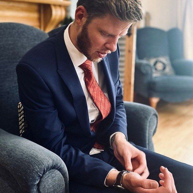 Wade John Beckett A photo of Wade John Beckett wearing a blue suit and red tie adjusting his watch whilst sitting in a blue wingback chair.